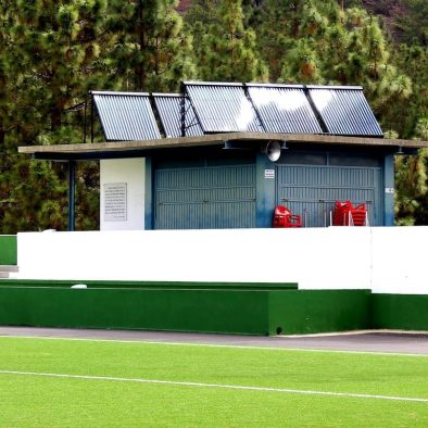 colectores solares en techo de módulo de servicios en instalación deportiva. Césped artificial en primer término. Al fondo módulo y árboles