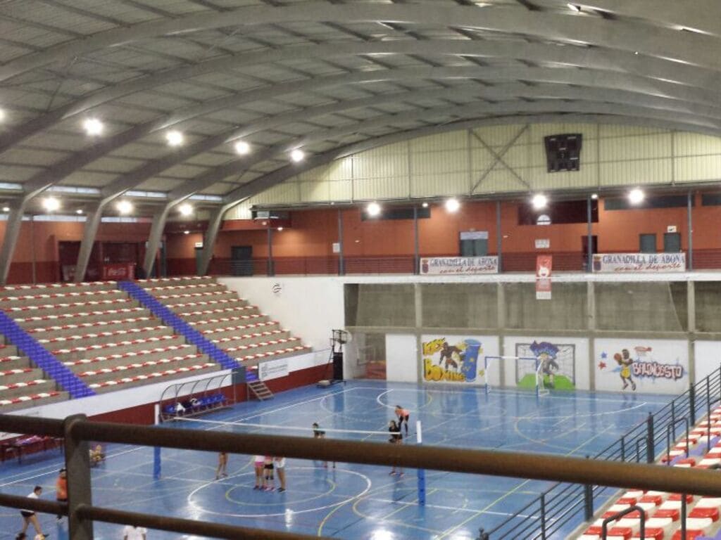 interior de pabellón de derpotes. Techo curvo gris, gradas de unas 10 filas en lateral izquierdo. Personas haciendo deporte sobre parqué azul