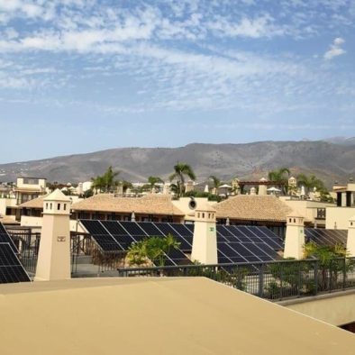 paneles solares en techo de conjunto de apartamentos. Chimeneas en primer término. Color construcción beige. Al fondo cielo azul y montañas