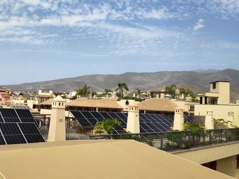 paneles solares en techo de conjunto de apartamentos. Chimeneas en primer término. Color construcción beige. Al fondo cielo azul y montañas