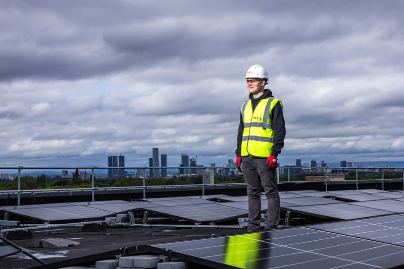 foto de operario de mantenimiento de paneles solares. Está de pie, con casco blanco y chaleco amarillo. Al fondo, cielo muy nuboso